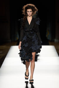 Model walking a runway in a black, ruffle-detailed dress with layered petal-like appliqués and minimalist sandals.