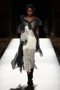Model walking runway in a sheer black over-lace dress with dramatic flutter sleeves and a white lace underdress, black boots.