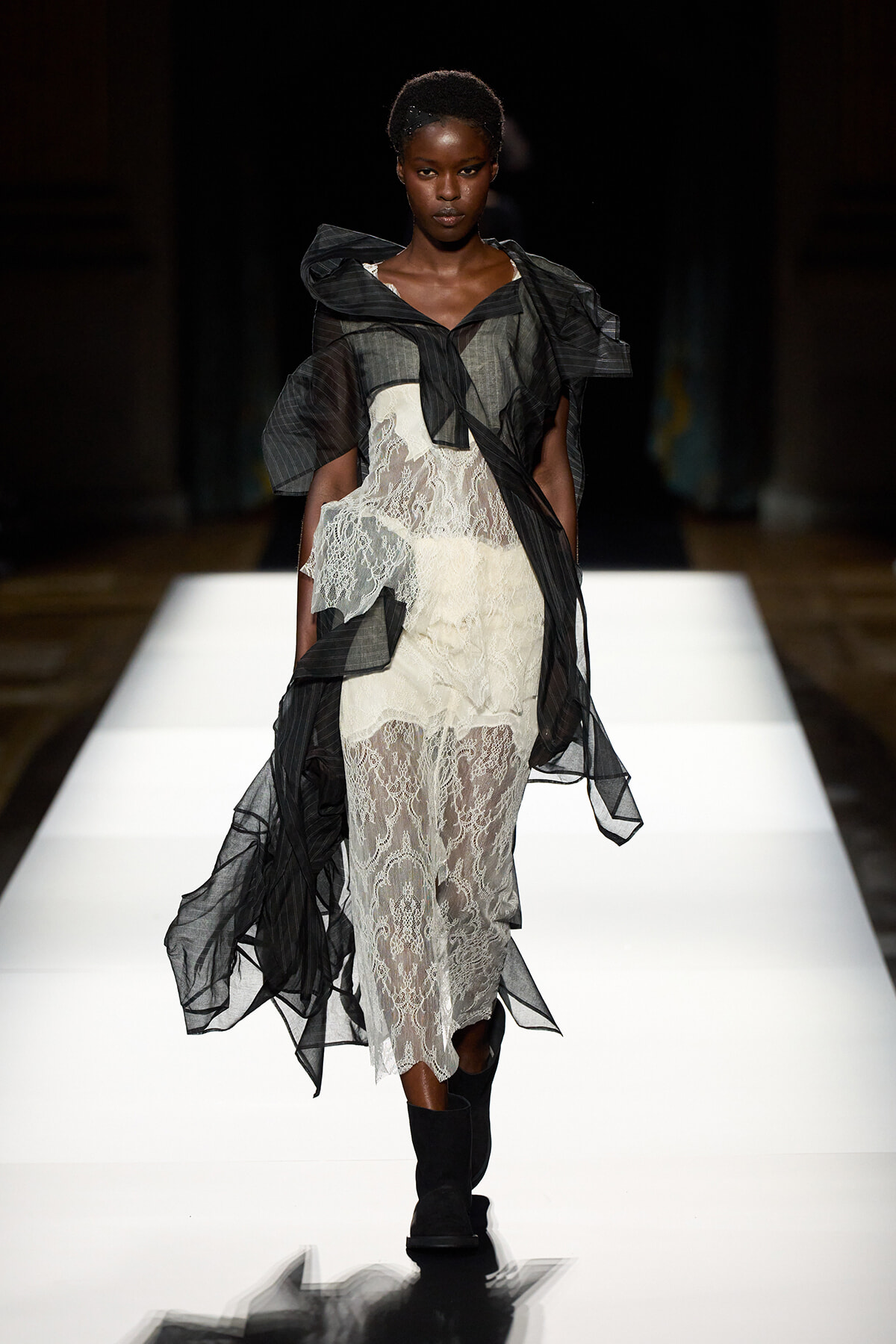 Model walking runway in a sheer black over-lace dress with dramatic flutter sleeves and a white lace underdress, black boots.