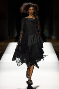 Black-clad model walks a runway in a voluminous, layered dress with lace sleeves and a braided side ponytail, on a white-lit catwalk.