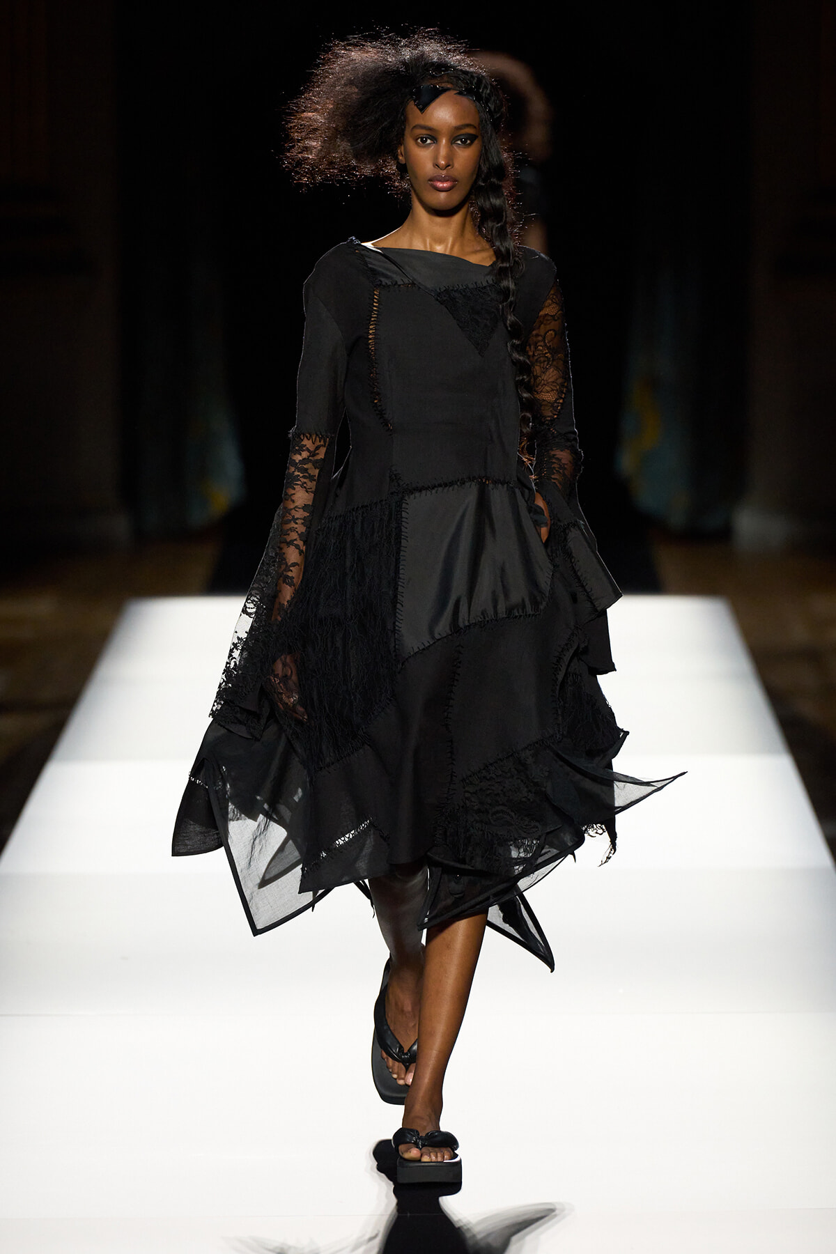 Black-clad model walks a runway in a voluminous, layered dress with lace sleeves and a braided side ponytail, on a white-lit catwalk.