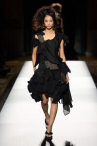 Female model walking a runway in a black sculptural, ruffled dress with sandals.