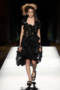 Model on a runway wearing a black, layered ruffled dress with shiny metallic panels and black sandals on a white catwalk.