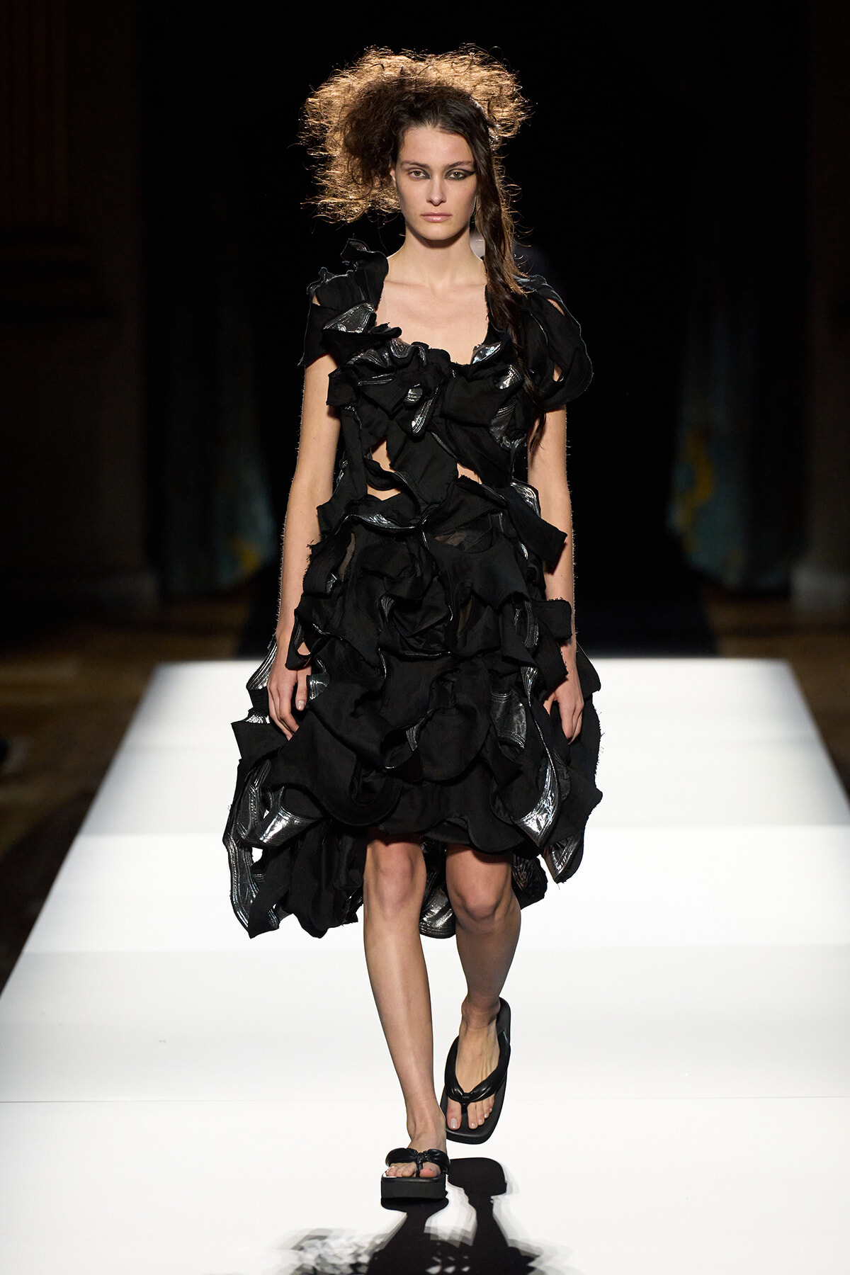 Model on a runway wearing a black, layered ruffled dress with shiny metallic panels and black sandals on a white catwalk.