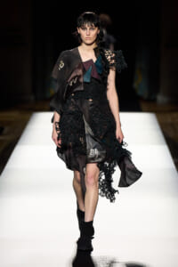 Model walks the runway in a black, lace-detailed dress with geometric satin panels and braided shoulder detail on a white catwalk.