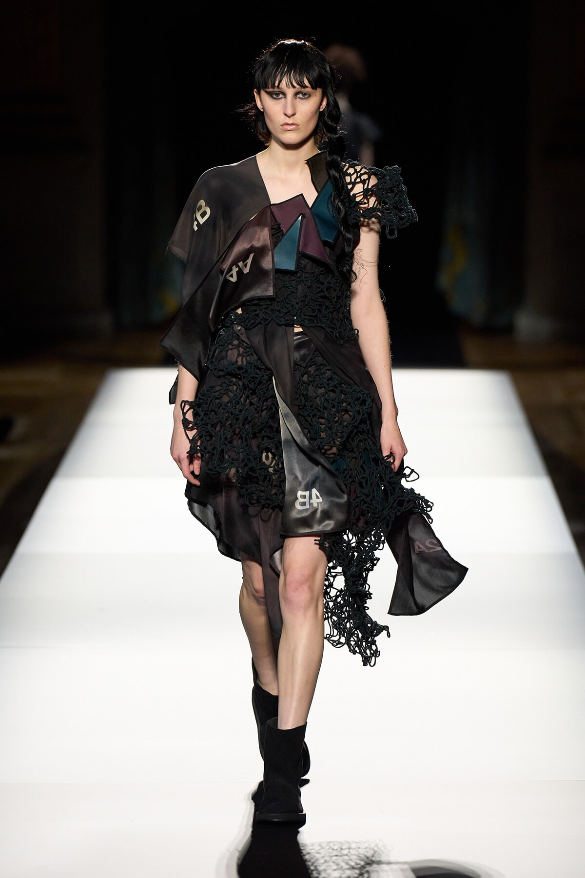 Model walks the runway in a black, lace-detailed dress with geometric satin panels and braided shoulder detail on a white catwalk.