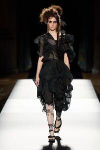 Runway model wearing a voluminous black ruffled sheer dress with bold makeup walking a white catwalk.
