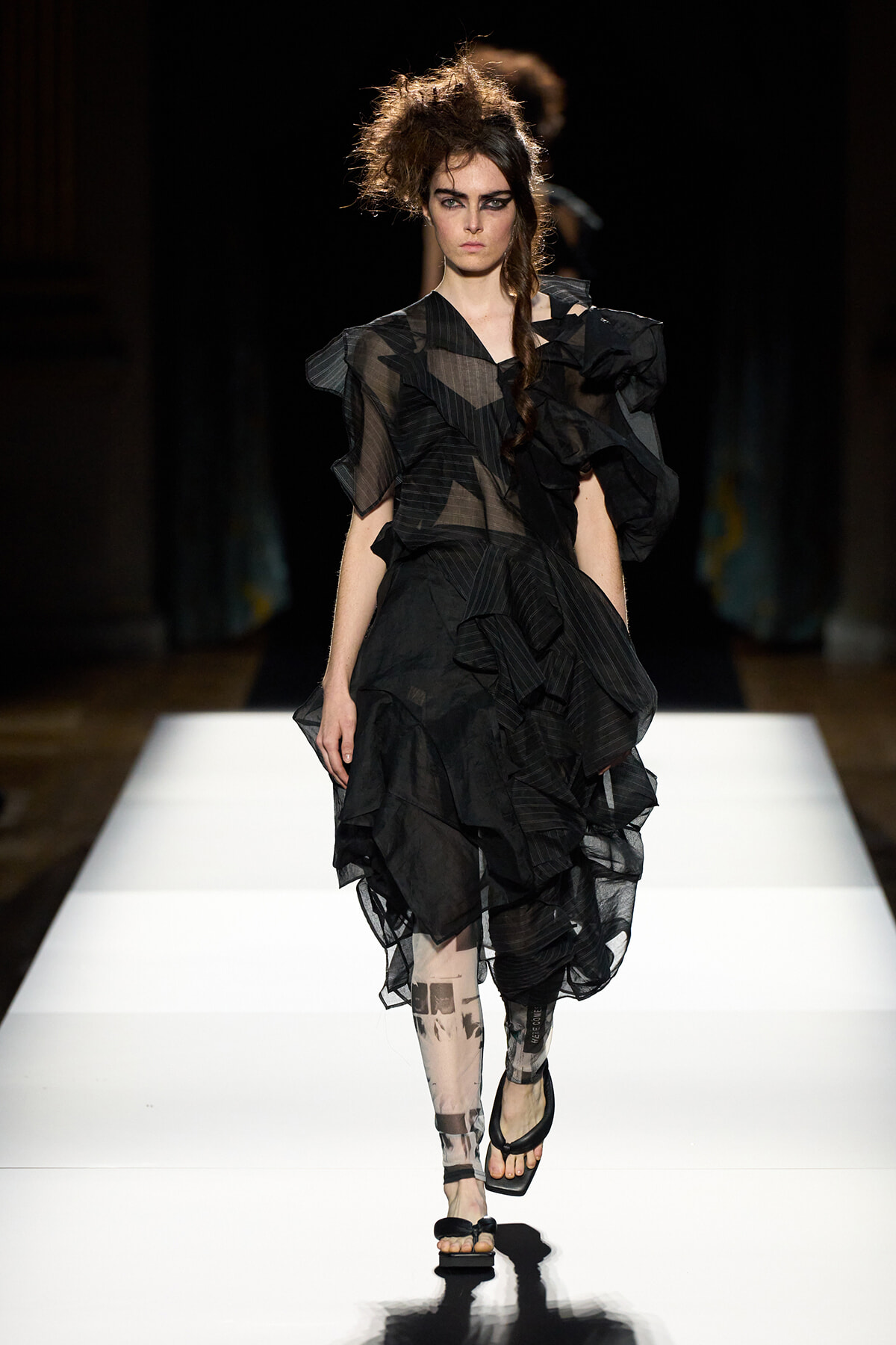 Runway model wearing a voluminous black ruffled sheer dress with bold makeup walking a white catwalk.