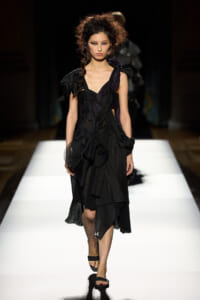 Model walks the runway in a black, layered dress with pleated panels and wrap details, wearing black sandals.