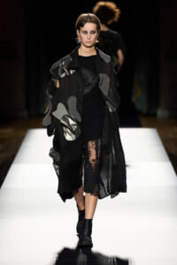 Model walking a fashion runway in a black oversized coat with gray patches and lace tights, black boots.