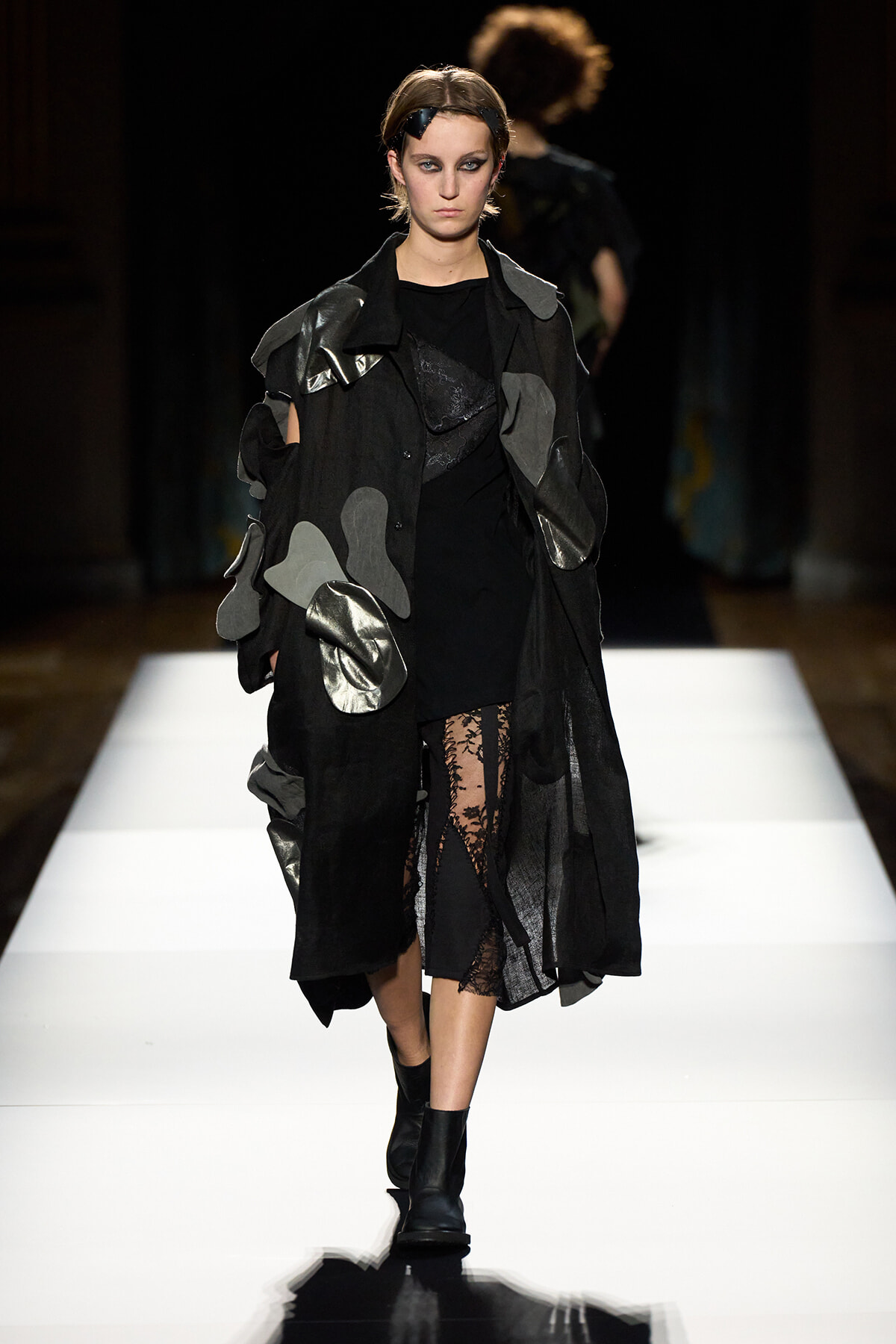 Model walking a fashion runway in a black oversized coat with gray patches and lace tights, black boots.