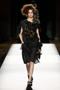 Female model walks a white runway wearing a black patchwork dress with oversized ruffles and olive-green accents.