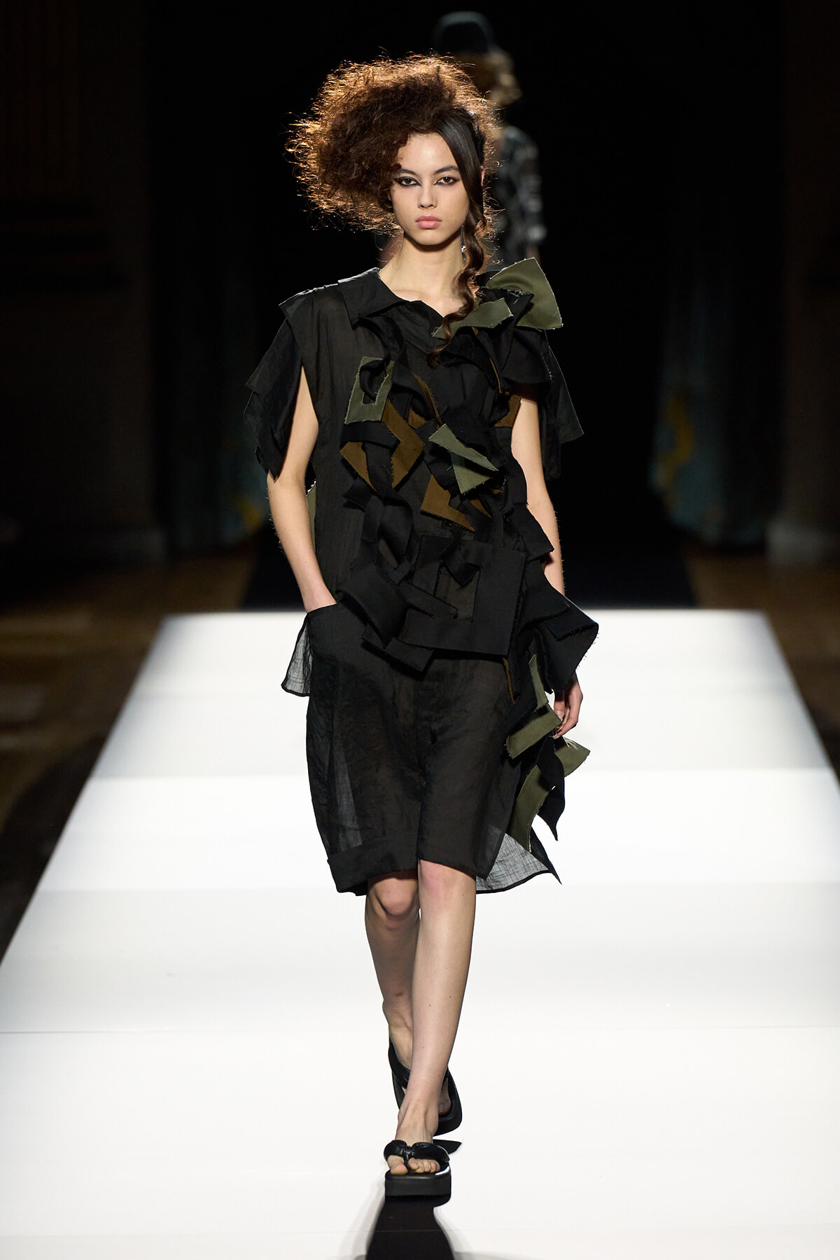 Female model walks a white runway wearing a black patchwork dress with oversized ruffles and olive-green accents.