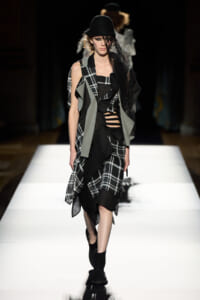 Model walking a fashion runway in a black hat, lace veil, and layered black-and-gray plaid ensemble with cutouts.