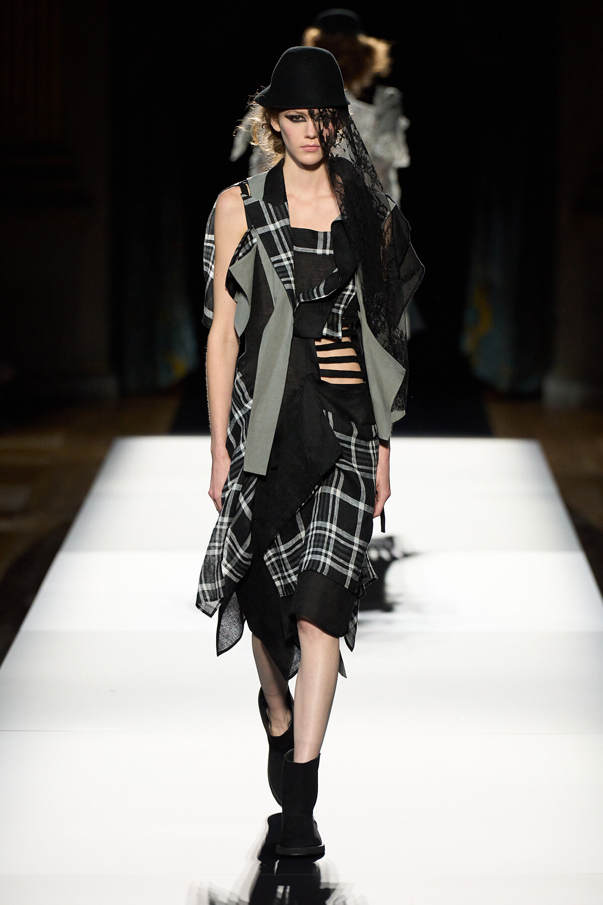 Model walking a fashion runway in a black hat, lace veil, and layered black-and-gray plaid ensemble with cutouts.