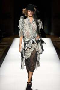 Model walks a fashion show runway in a sheer lace dress with ruffled sleeves and layered skirt, wearing a black hat and veil.