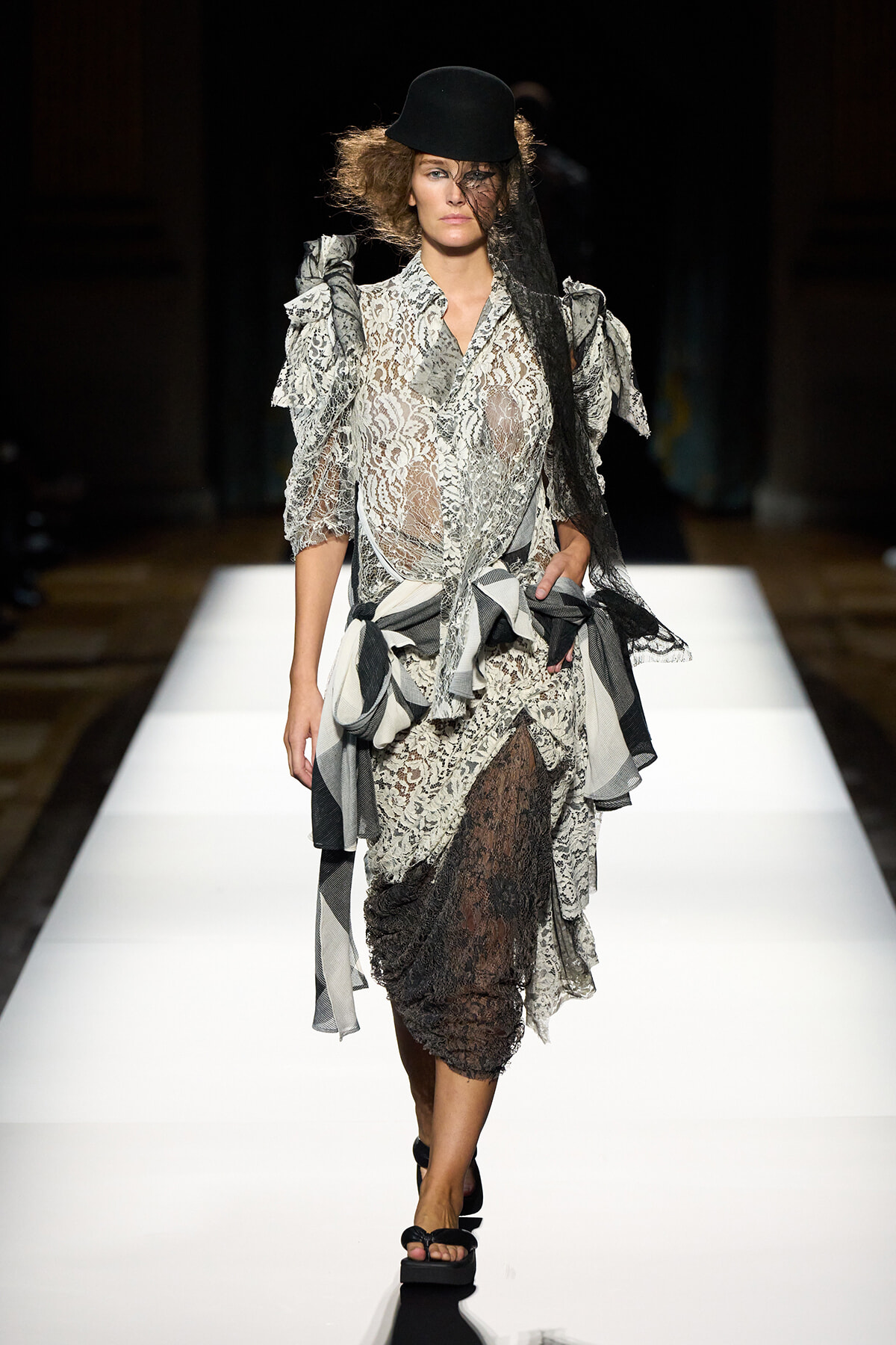 Model walks a fashion show runway in a sheer lace dress with ruffled sleeves and layered skirt, wearing a black hat and veil.
