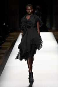 Model walks down a white runway wearing a black, layered dress with pleated ruffles and ankle boots.