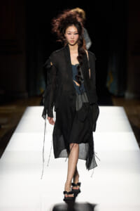 Runway model walking the catwalk in a sheer black layered coat over a dark outfit, with a voluminous updo hairstyle.