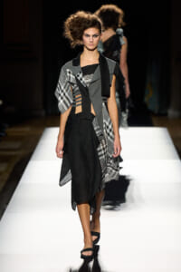 Model walks the runway in a black asymmetrical dress with gray panels, plaid accents, and a draped scarf. That’s a concise description of the look and context on the catwalk.