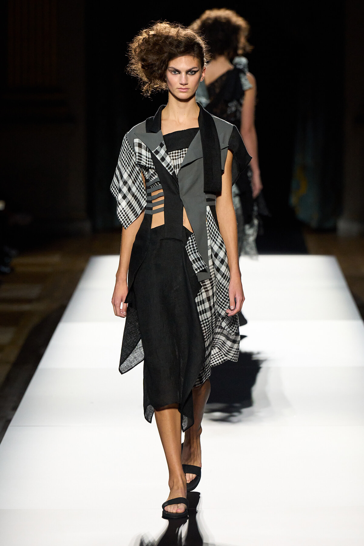 Model walks the runway in a black asymmetrical dress with gray panels, plaid accents, and a draped scarf. That’s a concise description of the look and context on the catwalk.