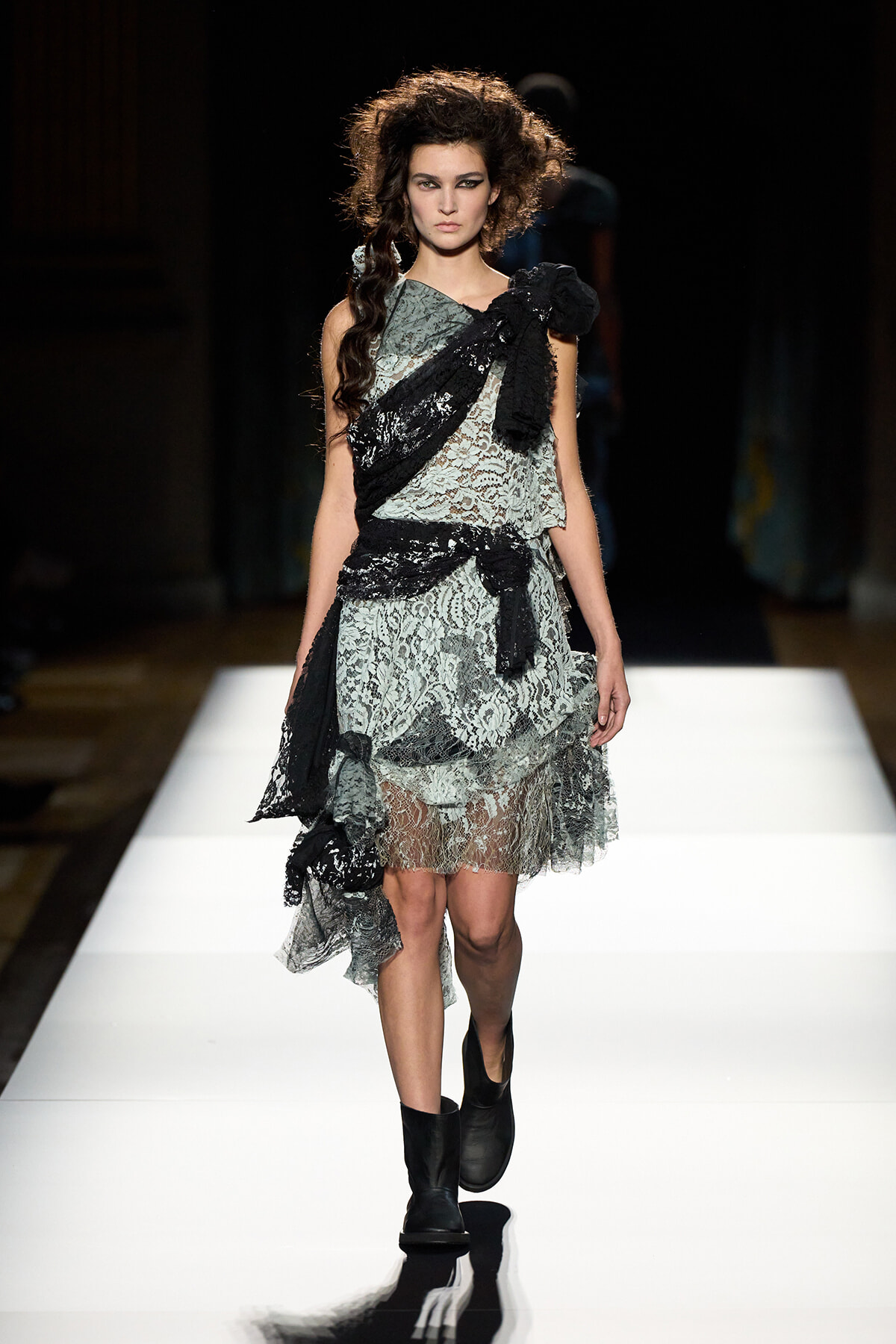 Female model walks a fashion runway in a gray lace, knee-length dress wrapped with black fabric and boots.