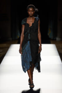Model walking a runway in a black and teal draped dress with ruffles and lace accents, paired with flat sandals.
