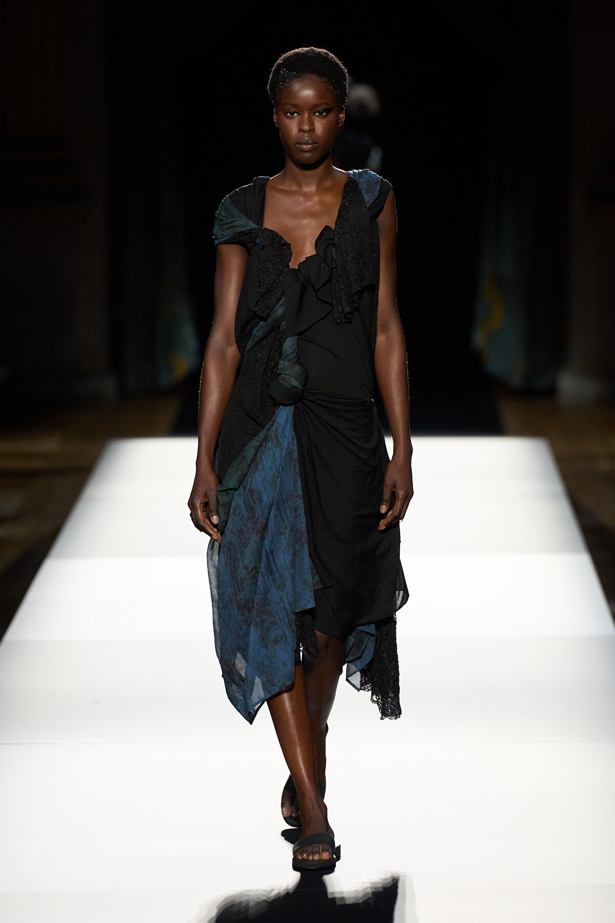 Model walking a runway in a black and teal draped dress with ruffles and lace accents, paired with flat sandals.