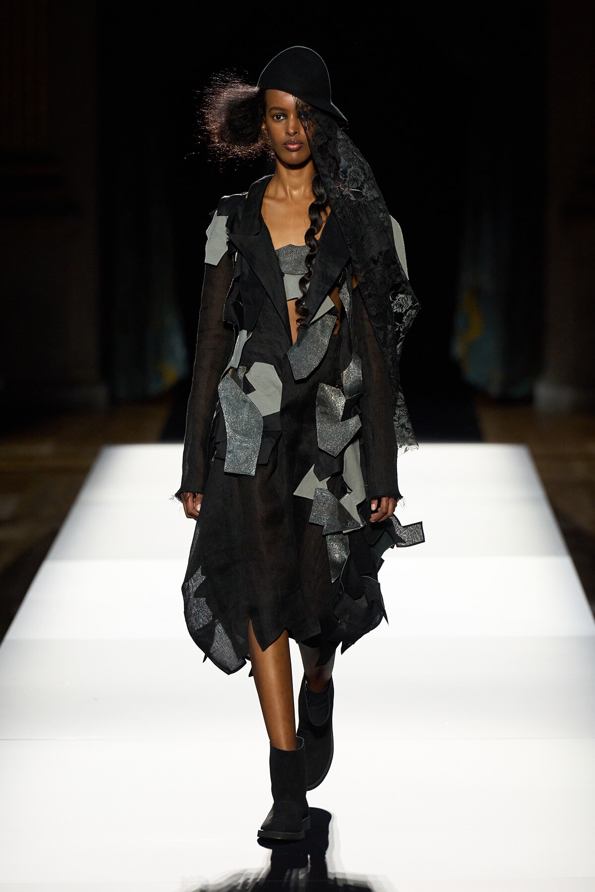 Fashion model walking a runway in a black patchwork coat with layered fabric scraps, wide-brim hat, and ankle boots.