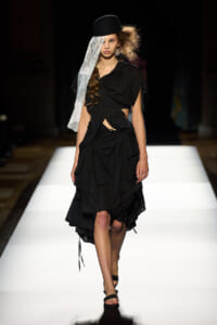 Model on a white runway wearing a black draped dress with asymmetrical folds, a black hat, and a sheer lace veil.