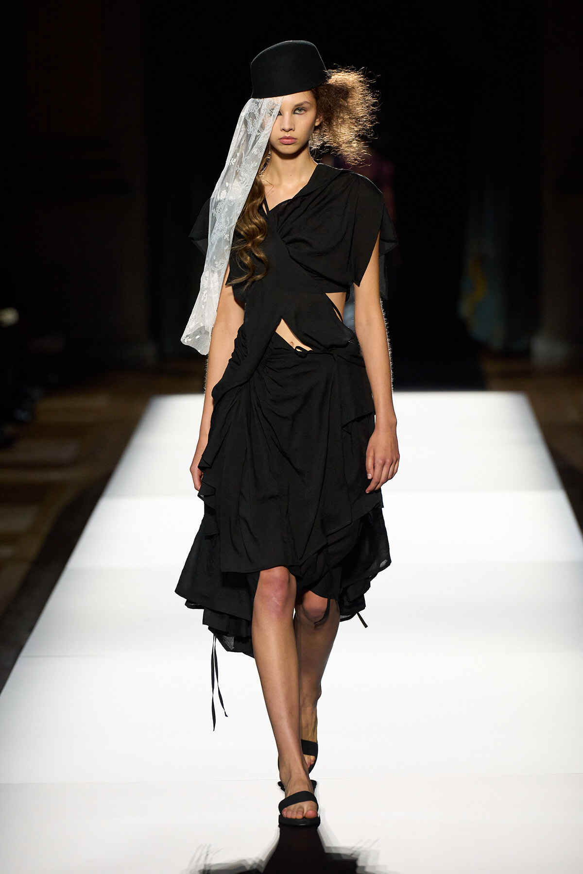 Model on a white runway wearing a black draped dress with asymmetrical folds, a black hat, and a sheer lace veil.