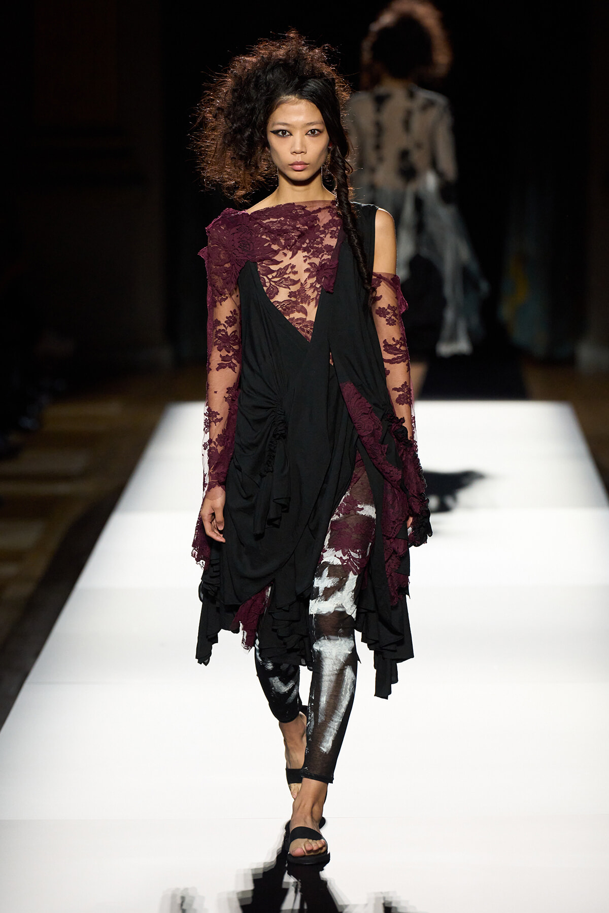 Model walks down a white runway in a black, draped dress with burgundy lace detailing and sheer sleeves, accessorized with black sandals.