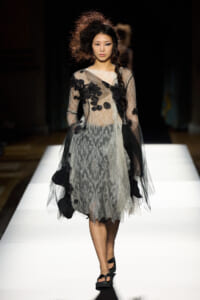 Model walks the runway in a sheer lace dress with black floral appliqués and a long braid, wearing black platform sandals.