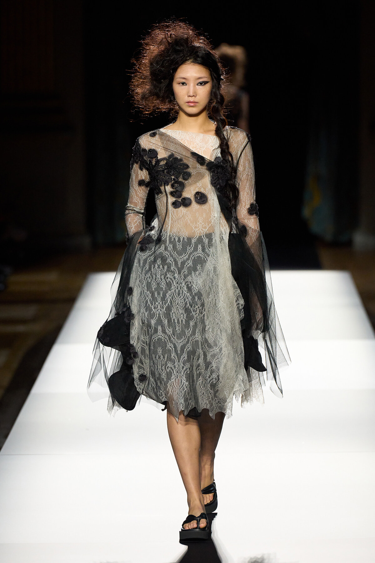 Model walks the runway in a sheer lace dress with black floral appliqués and a long braid, wearing black platform sandals.