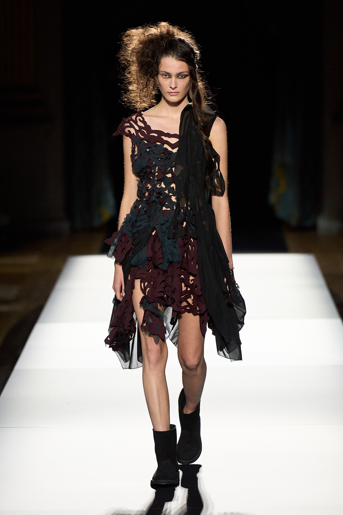 Model walks the runway in a black and burgundy laser-cut dress with sheer panels and black ankle boots on a white catwalk.