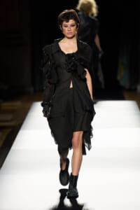 Model on a fashion runway wearing a black, layered dress with pleated, ruffled sleeves and chunky boots on a bright catwalk.