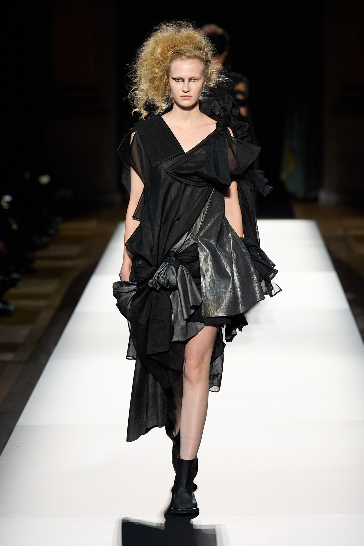 Model walks the runway in a black asymmetric dress with layered ruffles and sheer panels.