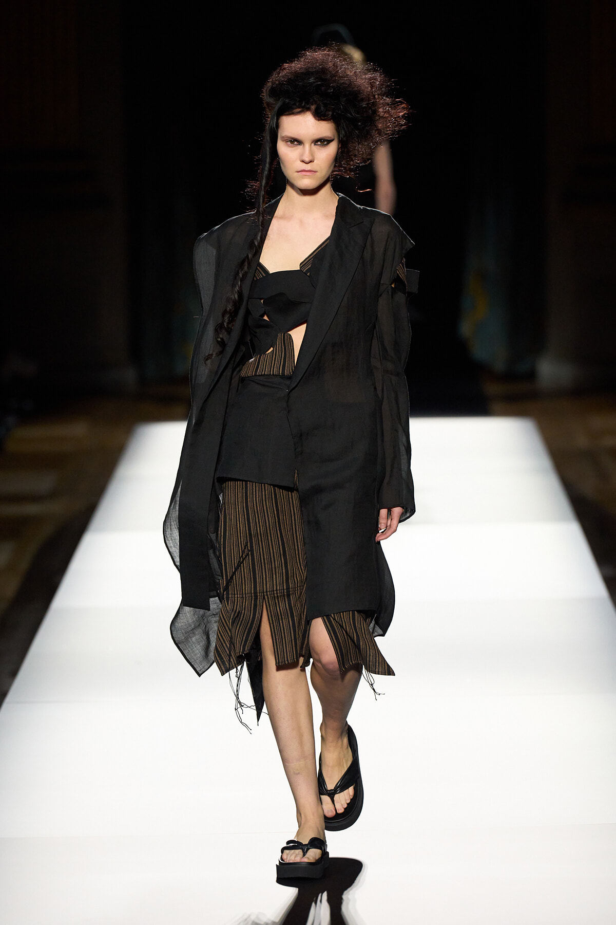 Model walking a runway in a black layered outfit with a sheer coat and striped skirt, minimalist styling.