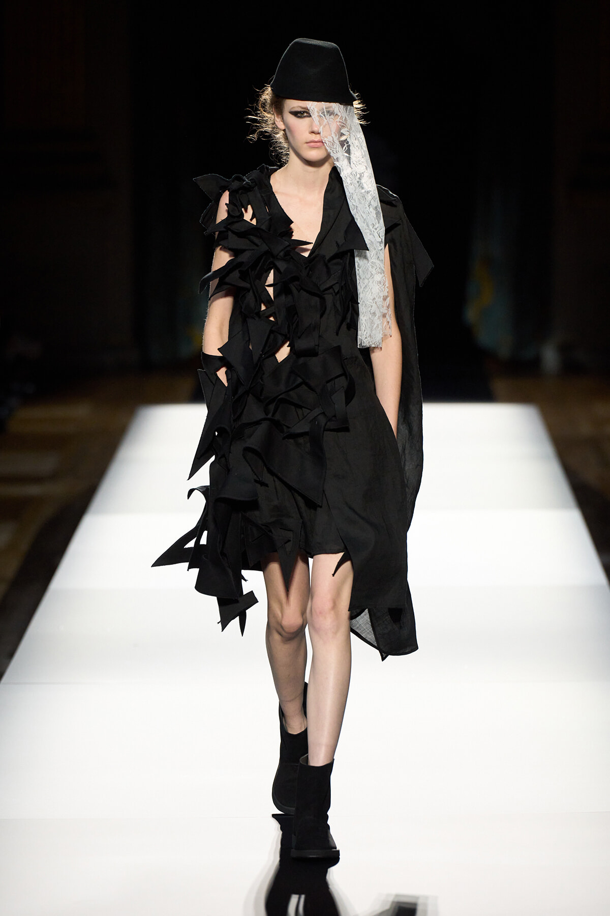 Model walking a runway in a black avant-garde dress with layered ruffles, paired with a black hat and lace veil.