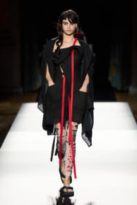 Model walking a runway in a black layered outfit with red ribbon accents and floral leggings, black sandals under bright white catwalk lighting.