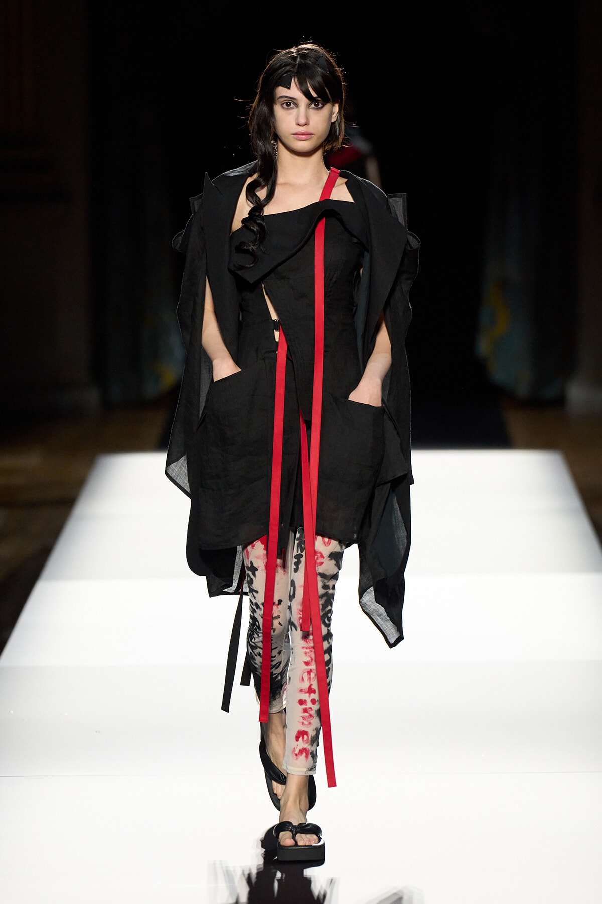 Model walking a runway in a black layered outfit with red ribbon accents and floral leggings, black sandals under bright white catwalk lighting.