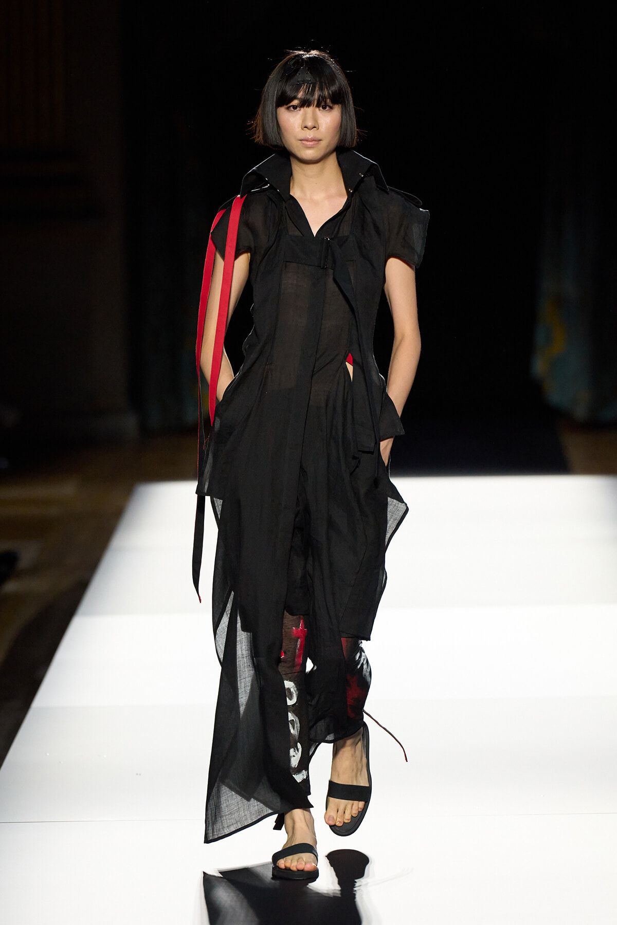 Model walks a fashion runway in a sheer, layered black dress with red straps and black sandals.