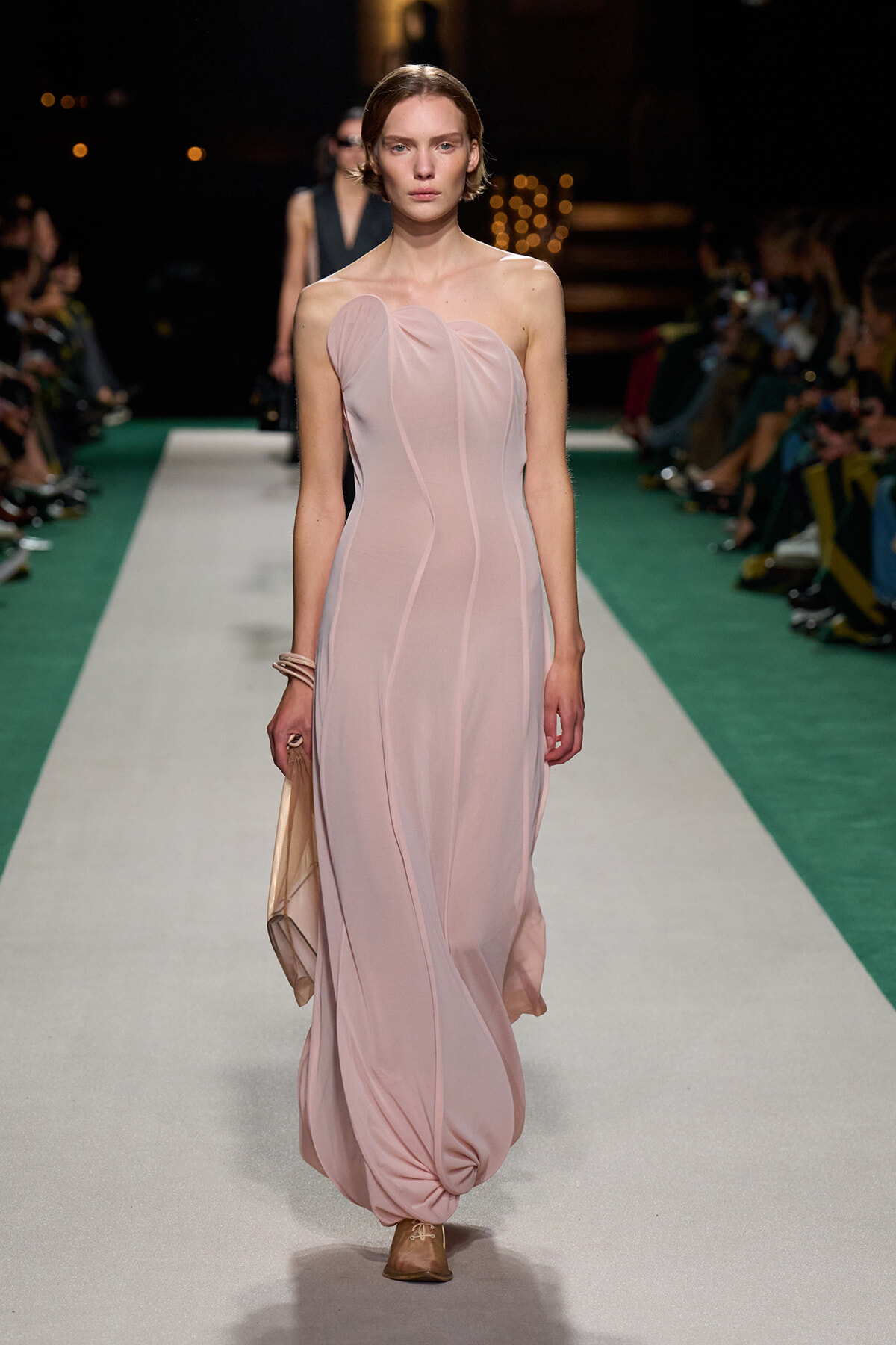 Model walks a fashion runway in a pale pink, one-shoulder draped gown with vertical seams, carrying a beige clutch and wearing brown flats as she glides down the white-green catwalk.