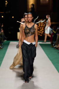 Model walking a fashion runway in a camo-drawn sheer top, black wide-leg pants, and a white belt; confident stride with a green and white runway behind.
