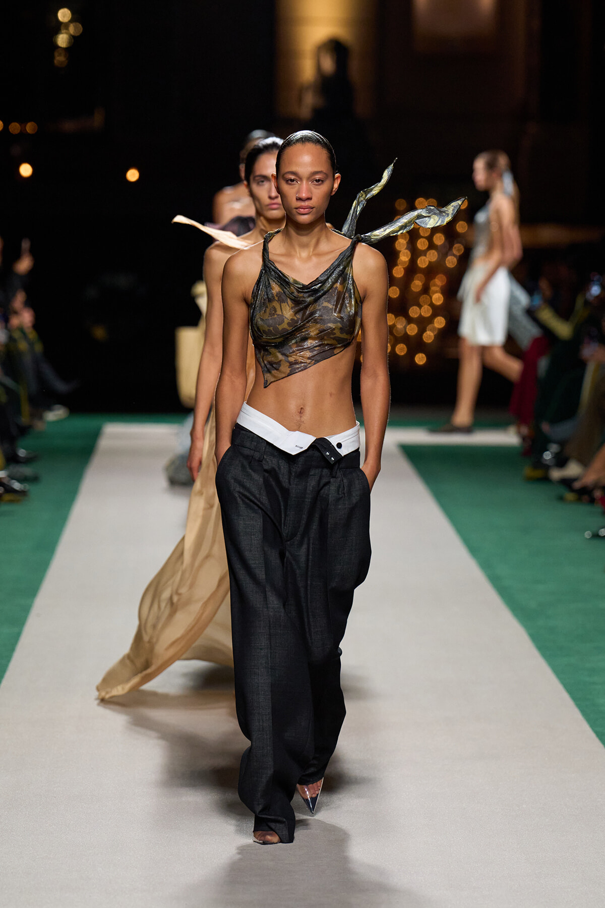 Model walking a fashion runway in a camo-drawn sheer top, black wide-leg pants, and a white belt; confident stride with a green and white runway behind.