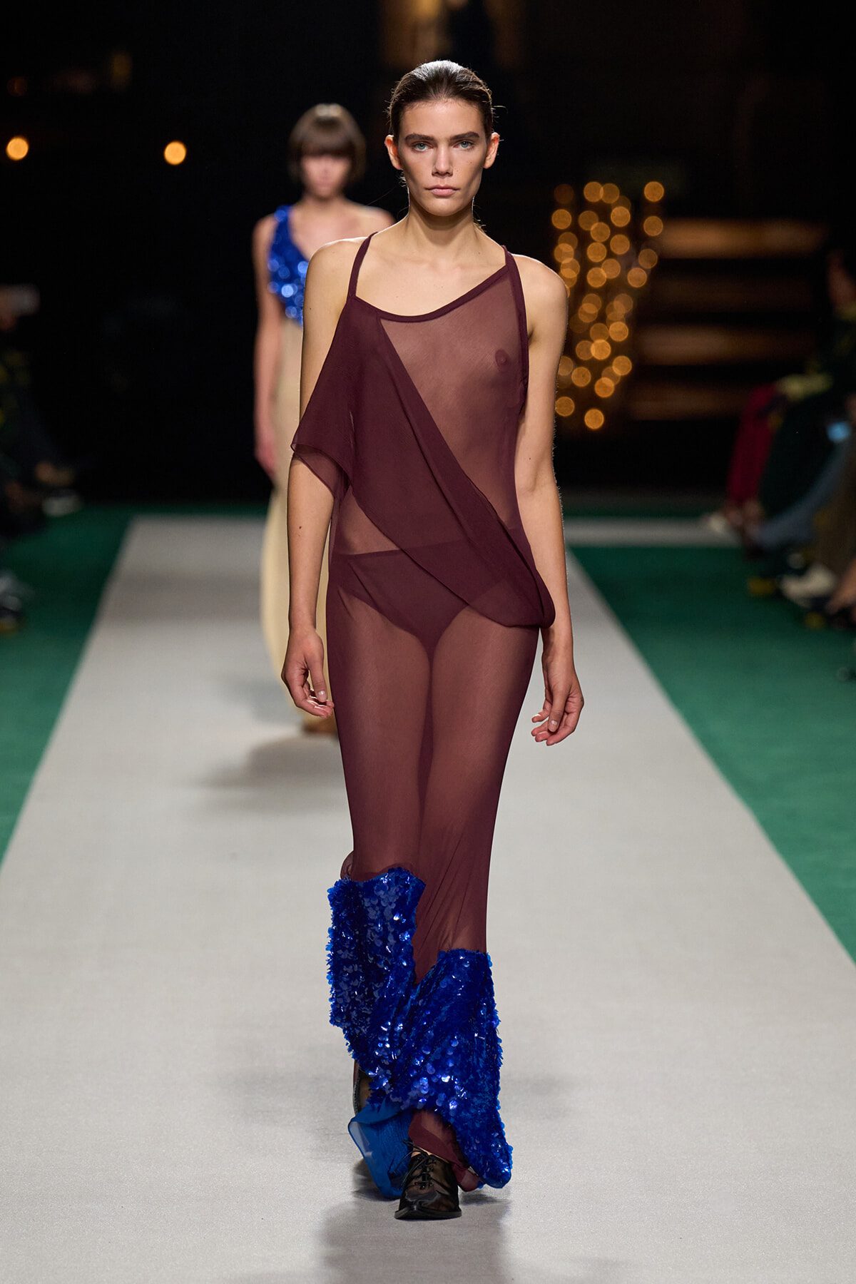 Model walks the runway in a burgundy sheer one-shoulder jumpsuit with blue sequin boots, spotlighted catwalk backdrop.