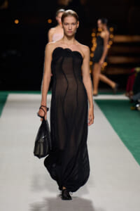Model walking a runway in a sheer, black strapless gown, carrying a black handbag at her side.
