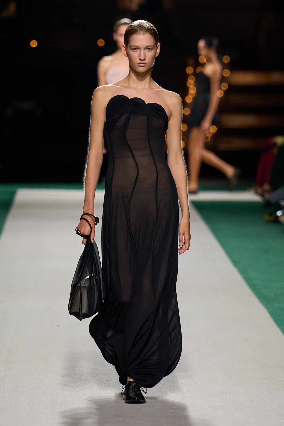Model walking a runway in a sheer, black strapless gown, carrying a black handbag at her side.