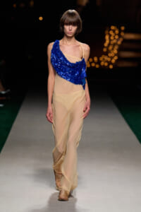 Model walks the runway in a sheer beige gown with a blue sequin drape over one shoulder, mid-stride on a lit runway at night