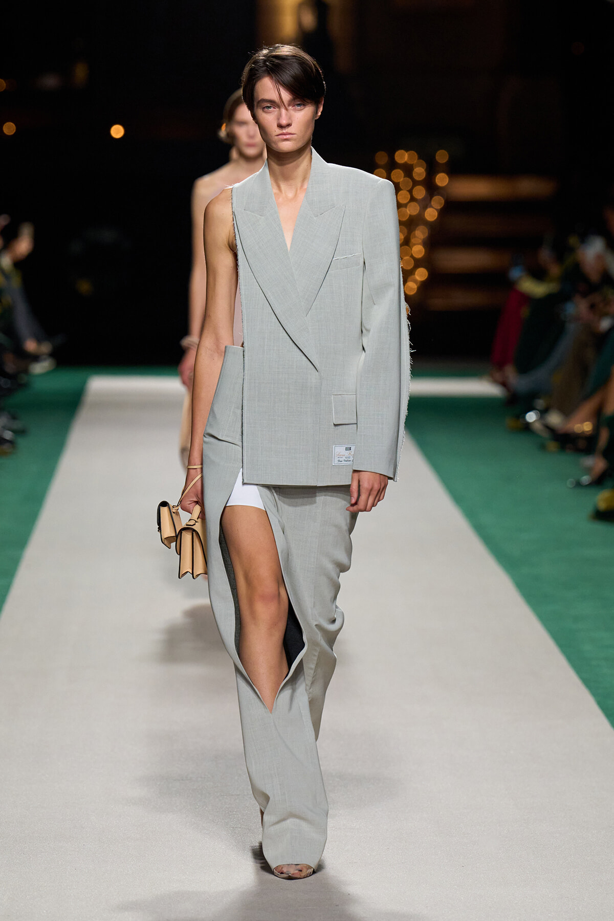 Model walks the runway in a light gray, oversized blazer with a high slit and matching wide-leg pants; she carries a beige handbag.